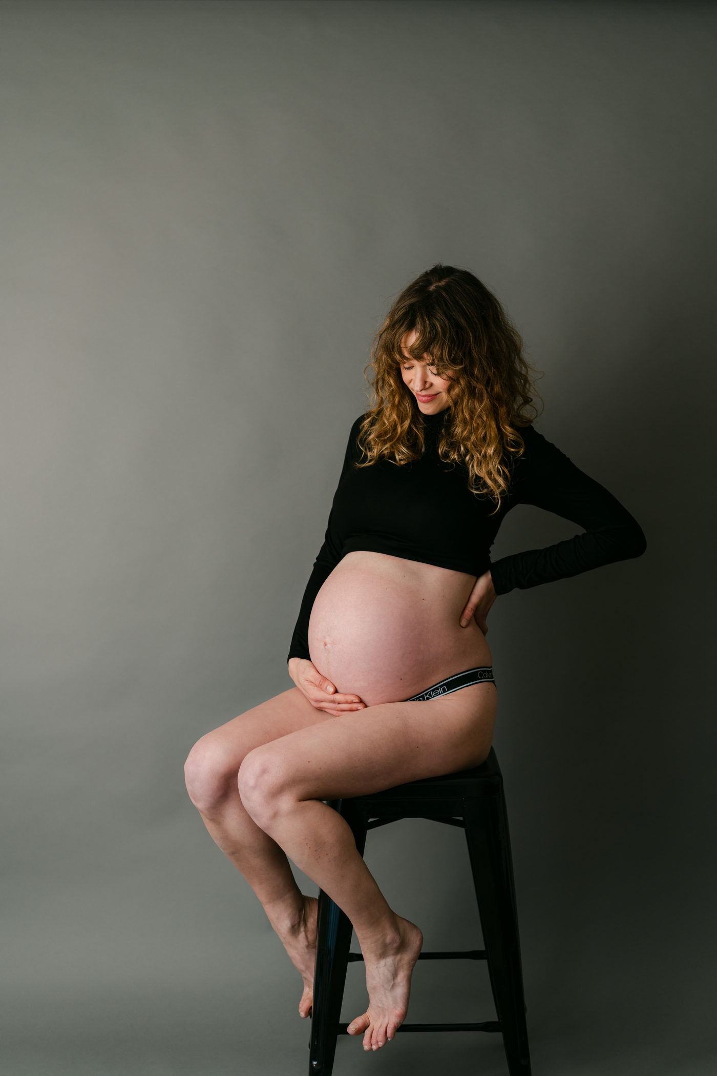 Mom sitting on stool looking at bump during Asheville, NC studio maternity session. Photo by Lauren Sosler Photography