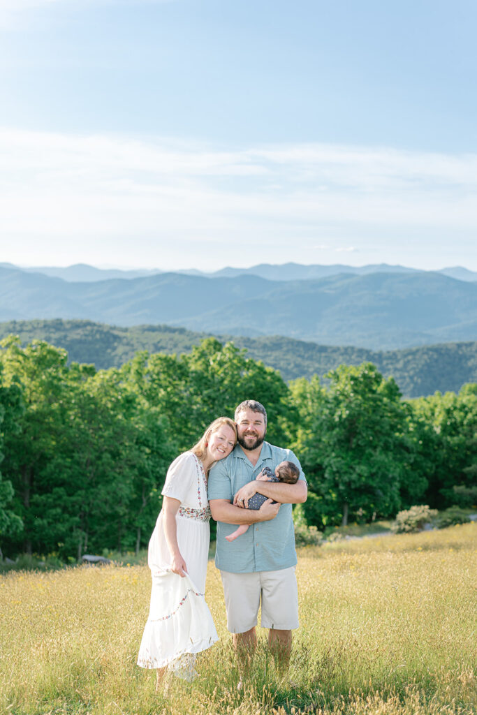 asheville photographer in the blue ridge mountains with parents and a baby. They are standing in a field, holding their baby, with mountains in the backdrop. 