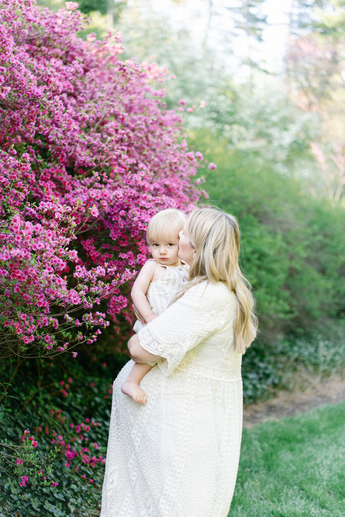 pregnant mother holding her toddler and kissing his forehead with a purple flowering tree behind them and asheville photographer documenting them.